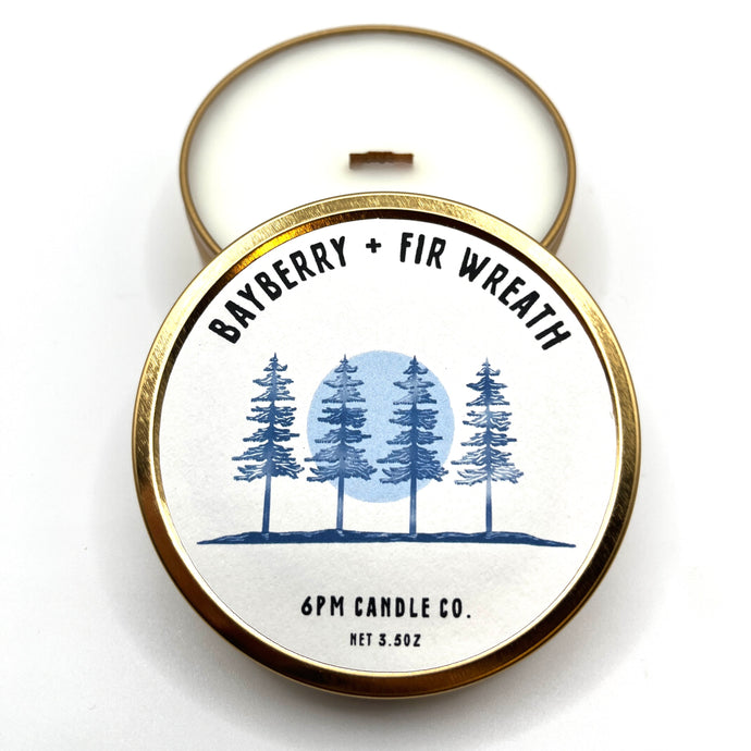 A travel-sized soy wax candle with a wooden wick, closed in a golden tin with a design featuring pine trees and a full moon. The candle is labeled 'Bayberry + Fir Wreath' by 6pm Candle Co.