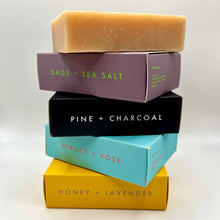 Load image into Gallery viewer, Stacked bar soaps in various boxed packages, each with different labels such as Sage + Sea Salt, Pine + Charcoal, Spruce + Rose, Honey + Lavender, and Salt + Pepper.

