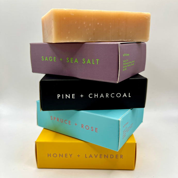 Stacked bar soaps in various boxed packages, each with different labels such as Sage + Sea Salt, Pine + Charcoal, Spruce + Rose, Honey + Lavender, and Salt + Pepper.