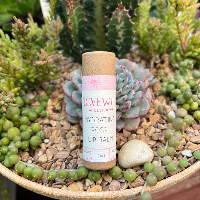 A tube of Lovewild Design Hydrating Rose Lip Balm placed on a round tray with various small plants and a large cactus in the background.